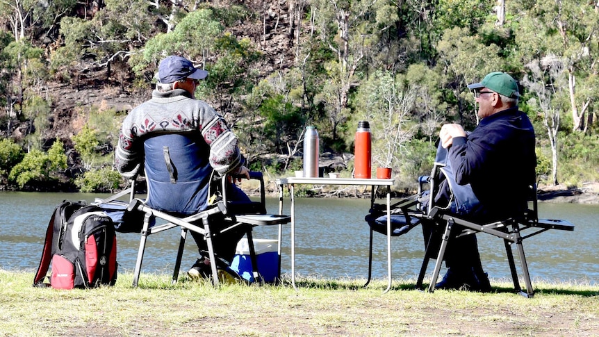 Two older men with grey hair sit on camping chairs by a lake.