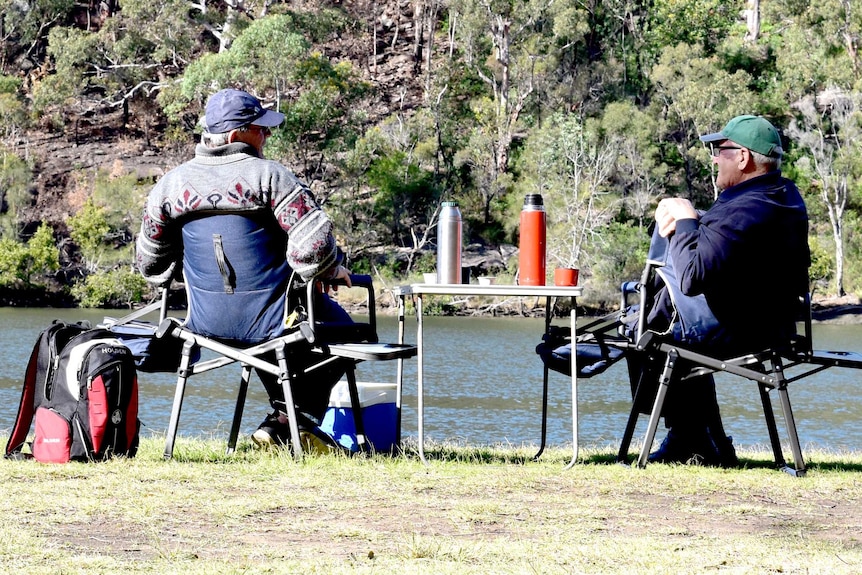 Two men in caps and jacket near a river talking as they sit on camping chairs and a table with a thermos.