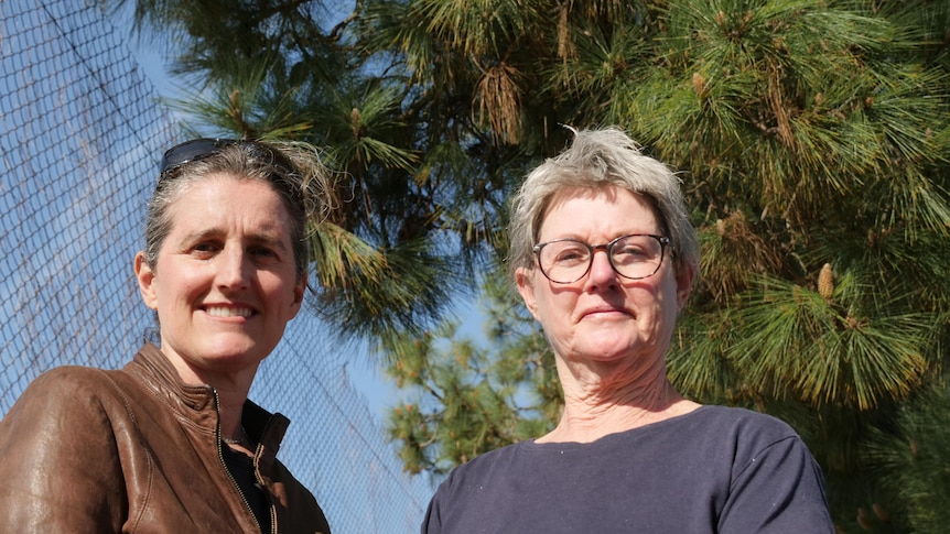 Two women look at the camera with a tennis fence and pine tree in the background