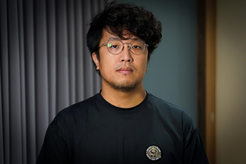 A young man wearing wire-rimmed glasses and a black T-shirt looks directly to camera with serious expression