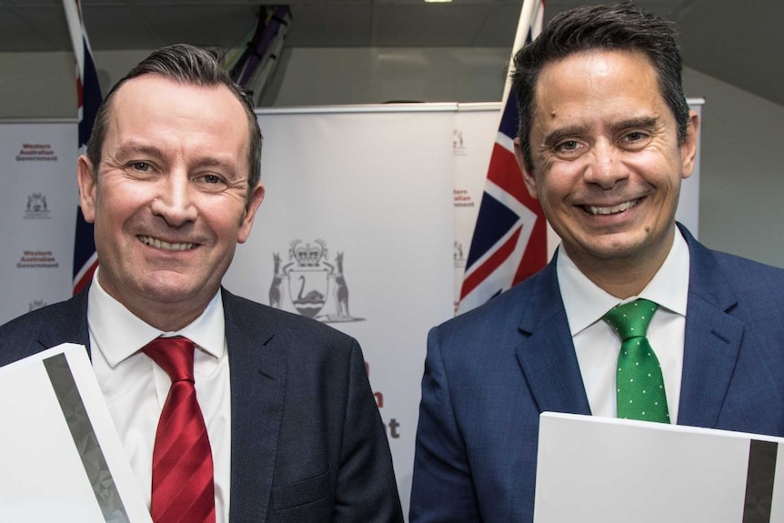 Mark McGowan and Ben Wyatt smile while holding the 2018 budget, in front of the Australian flag.