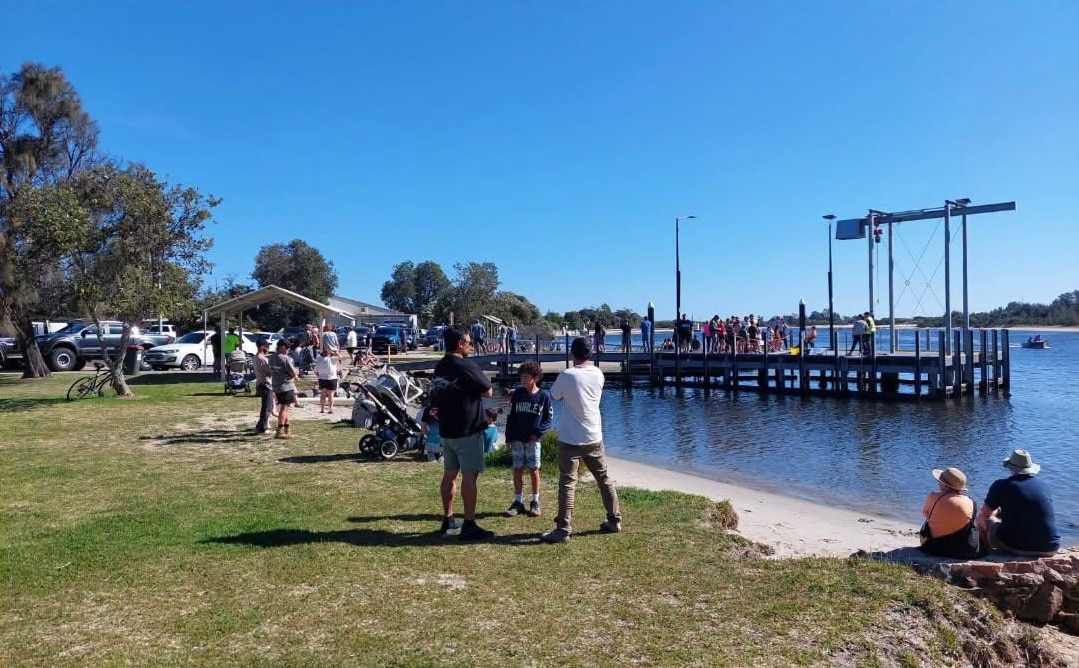 A crowd of people standing and watching on the grass and on a jetty, near water, small coastal town