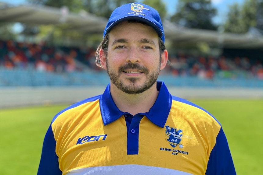 A man in a yellow and blue cricket uniform smiles at the camera.
