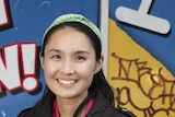 Alice Pung, with green headband, black jacket and wide smile, stands in front of a wall painted with bright advertising slogans.