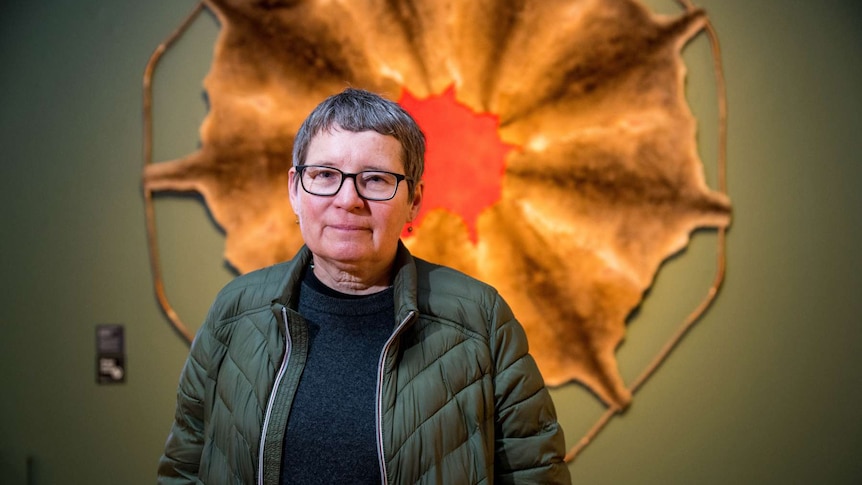 Image of woman in 50s with grey short hair standing in front of artwork made from kangaroo skin and red-dyed wool.