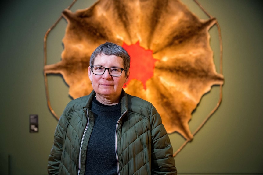 Image of woman in 50s with grey short hair standing in front of artwork made from kangaroo skin and red-dyed wool.