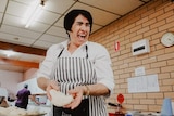 An older woman with dark black hair wearing an apron laughs as she handles a piece of dough in a brick-walled kitchen.