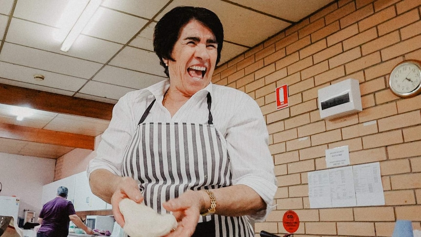 An older woman with dark black hair wearing an apron laughs as she handles a piece of dough in a brick-walled kitchen.