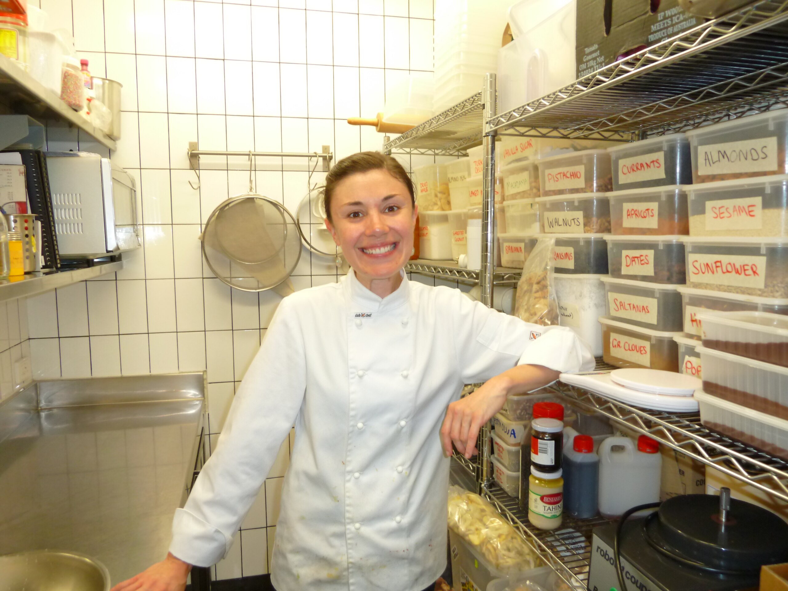 Woman in chef whites smiles standing in a commercial kitchen. Containers of ingredients on shelf beside