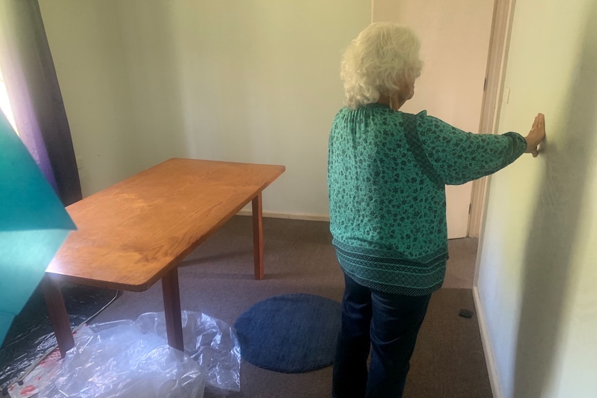 A photo taken from the back of a woman with grey hair, bright turquoise top, touching the wall, dining table is behind her.