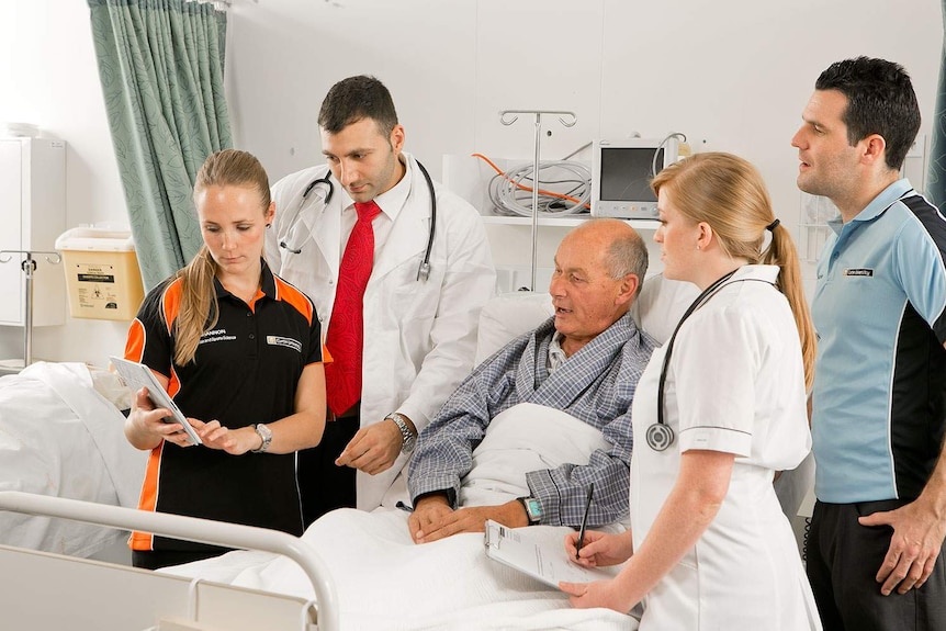 An older man sits up in a hospital bed as students and health professionals discuss treatment