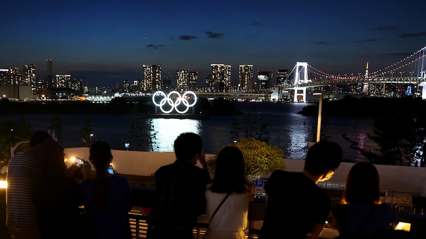 People look over a Tokyo skyline with a lit-up Olympic Rings in the background. 