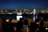 People look over a Tokyo skyline with a lit-up Olympic Rings in the background. 