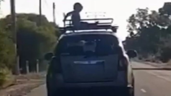 A grainy screenshot of a child sitting on a roof rack on the top of a car as it drives down a street.