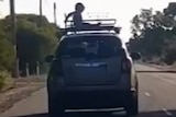 A grainy screenshot of a child sitting on a roof rack on the top of a car as it drives down a street.