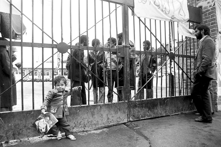 Child holding photo of the Ayatollah holds onto closed gates with group of men behind it.