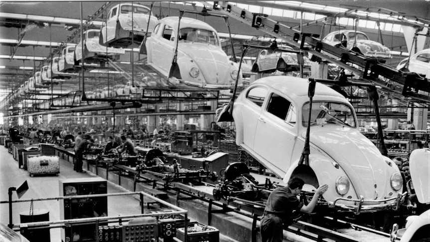 A black and white photo shows rows of Volkswagen Beetle chassis assembly lines.