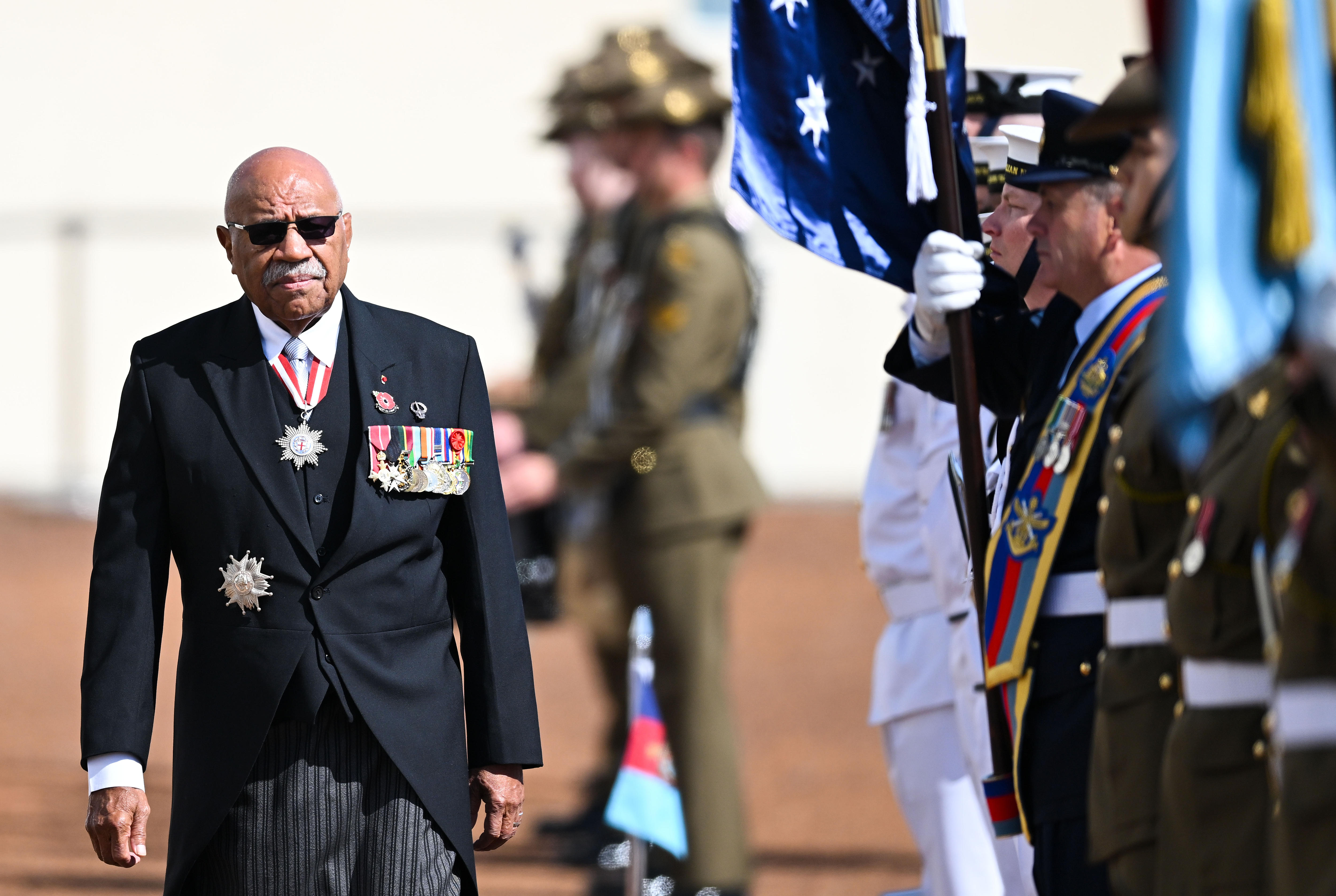 A man wearing a suit with military badges walks past a line of soldiers