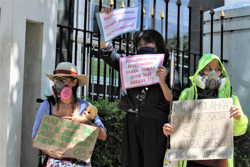 Three people holding signs, a doll, and wearing breathing tools.