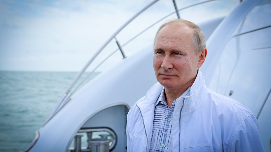 Vladimir Putin in a white member's only jacket standing on a yacht, with a slight smile on his face