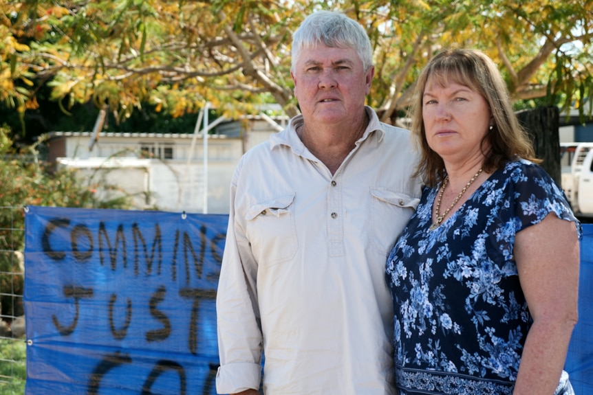 a man and woman in casual clothes stand in front of a leafy tree, with serious expressions on their face. 