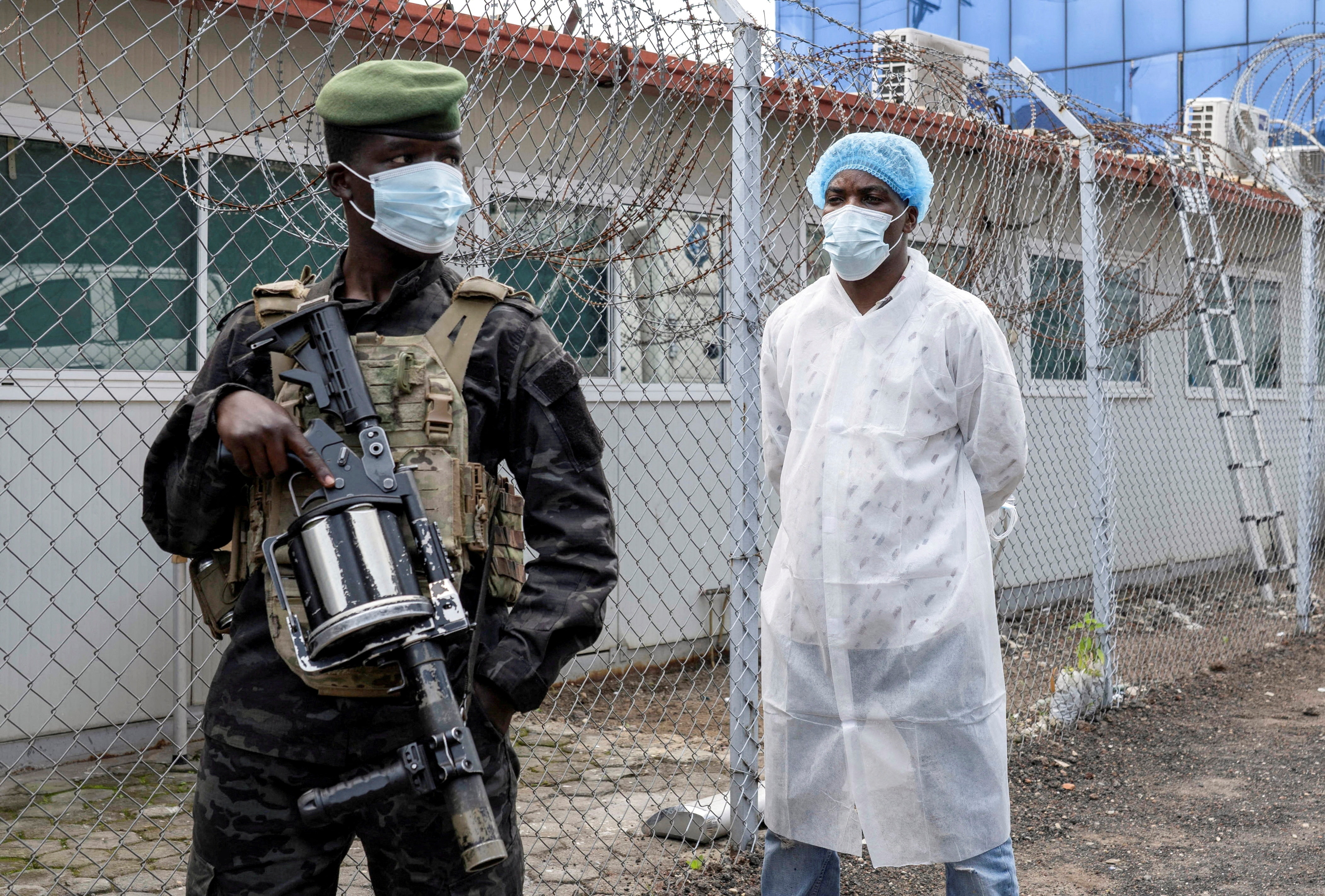 A man in military uniform standing guard in front of another in PPE.
