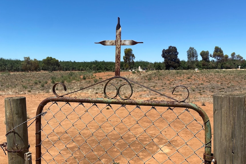 A rusty gate has a metal cross on the top, behind it is a green treeline with white gravestones in the distance