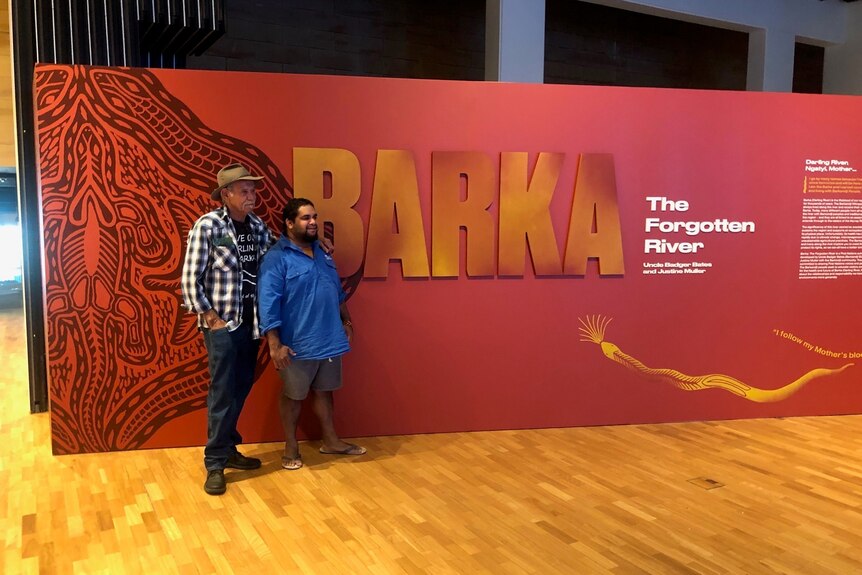 Two Aboriginal men, one wearing a hat standing in front of a red wall with writing on it in a museum.