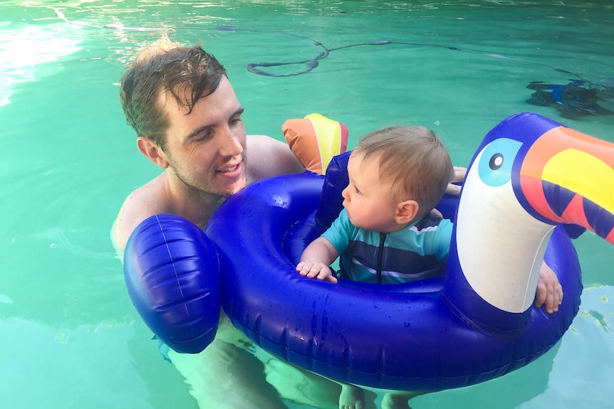 A father holds his baby who is sitting in a floating ring in the pool