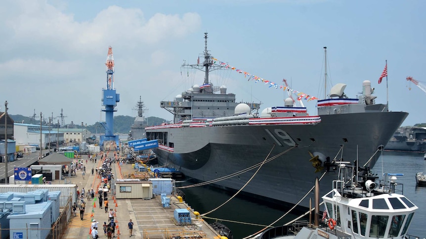 A large US Navy ship is docked and dressed with bunting in the US national colours beside a line of people to go aboard.