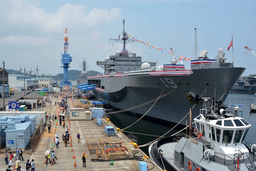 A large US Navy ship is docked and dressed with bunting in the US national colours beside a line of people to go aboard.