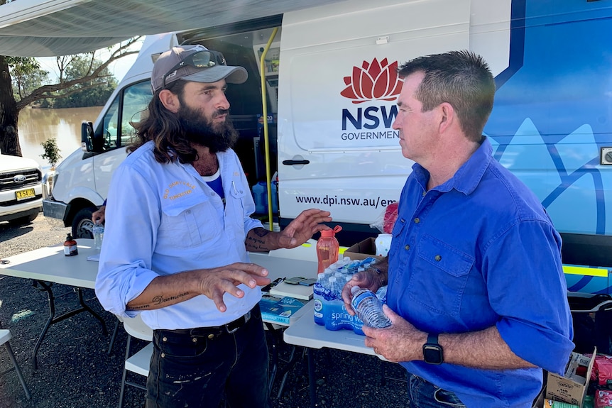 Two men in blue collared shirts speak to each other in front of a white and blue van.