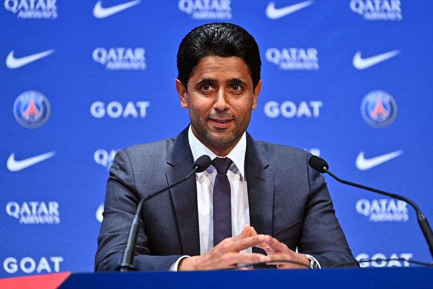 A man in a suit speaks at a media conference with a blue backdrop