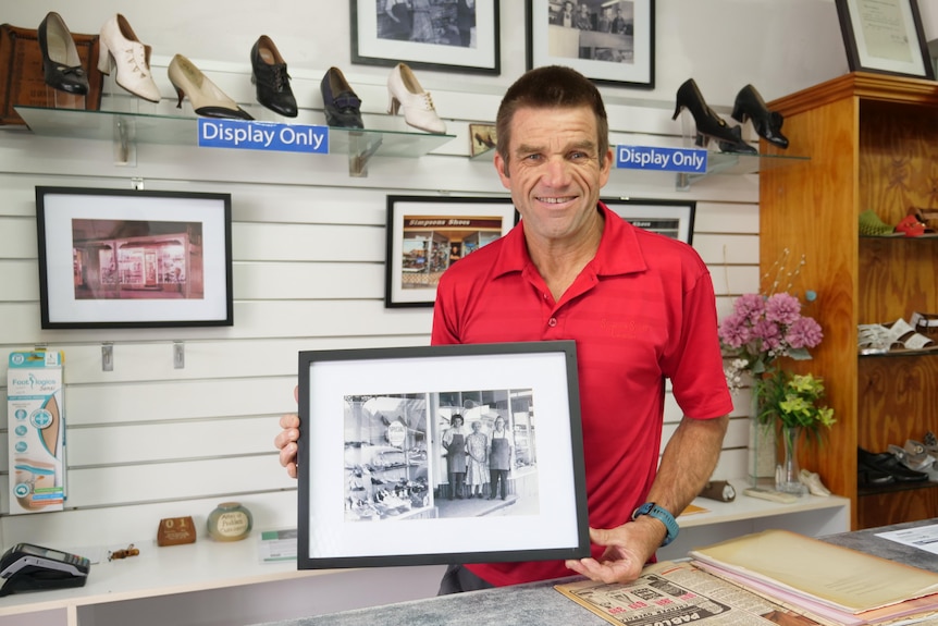 Craig Simpson holding a picture of his Pop, mother and father in front of a wall of shoes on display 