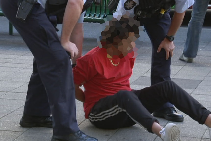 A young man sits on the ground with his hands behind his back. Two police officers stand beside him.