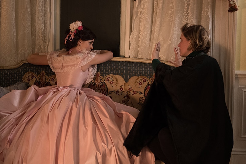 Emma Watson, in a large pink ball gown, slumps on a window seat while Greta Gerwig, not in costume, kneels and speaks to her