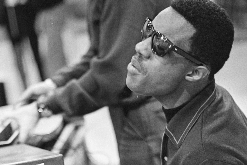 A monochrome shot of a young Stevie Wonder with glasses on playing a piano