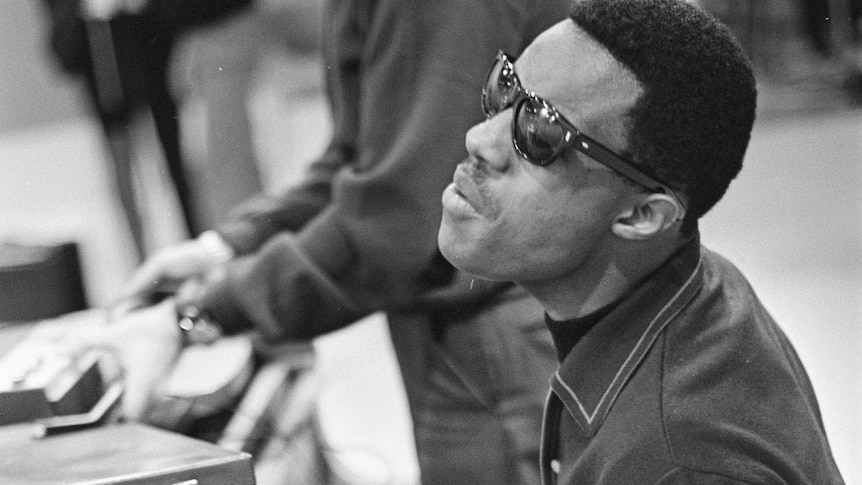 A monochrome shot of a young Stevie Wonder with glasses on playing a piano