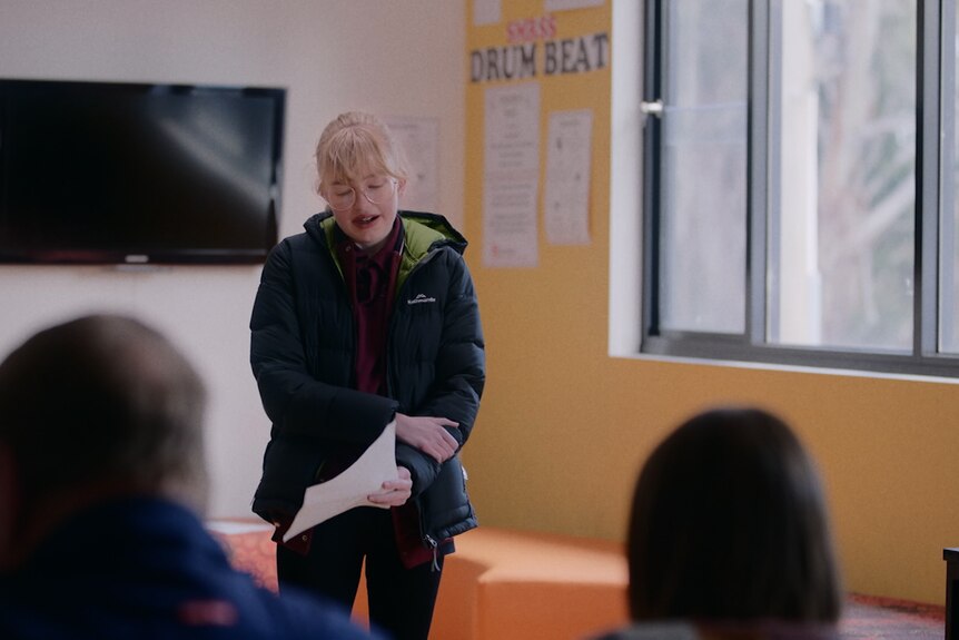 A teenage girl with blonde hair wearing a puffer jacket reads from a piece of paper during an audition.