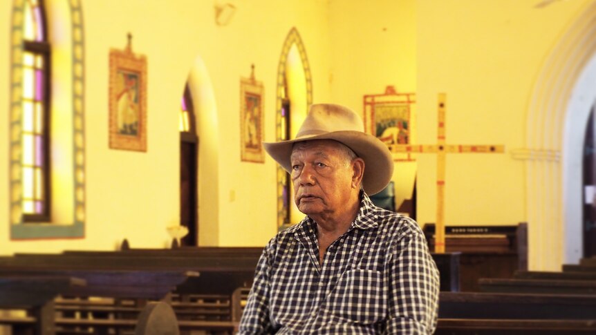 An old man, wearing a checkered shirt and farming hat, sitting alone in a church. There are tears in his eyes.