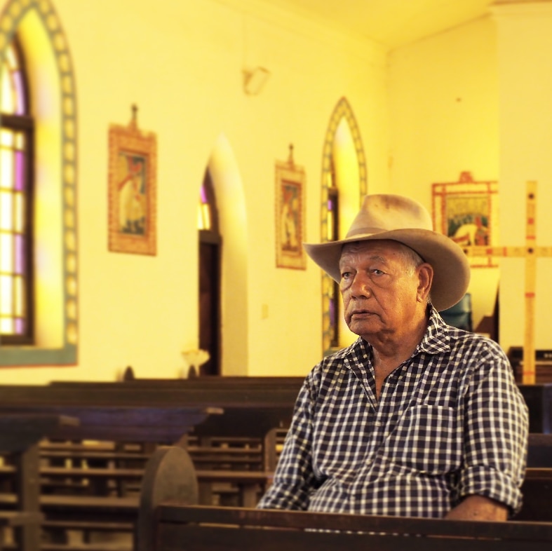 An old man, wearing a checkered shirt and farming hat, sitting alone in a church. There are tears in his eyes.