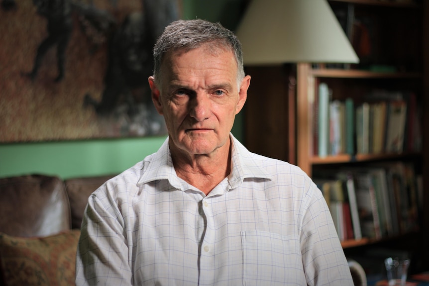 Andrew McDonald sitting in a room with a bookcase in the background looking at the camera, unsmiling