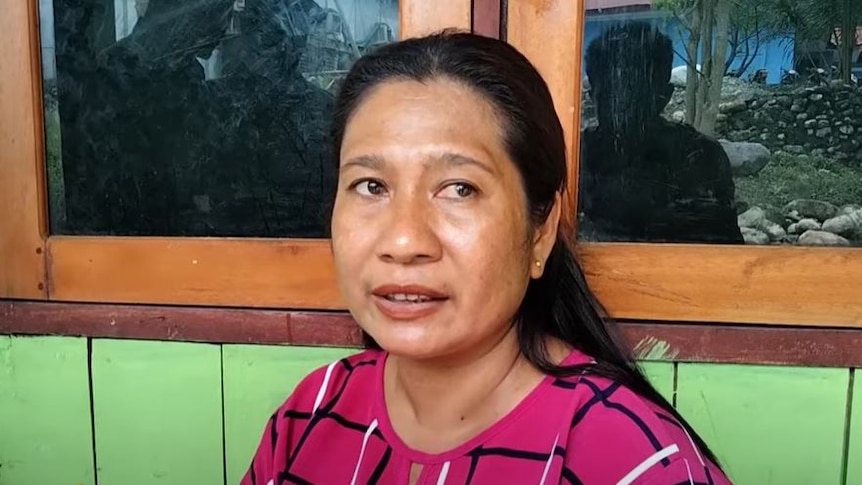 A Timorese woman in a pink shirt sits in front of a green wall 