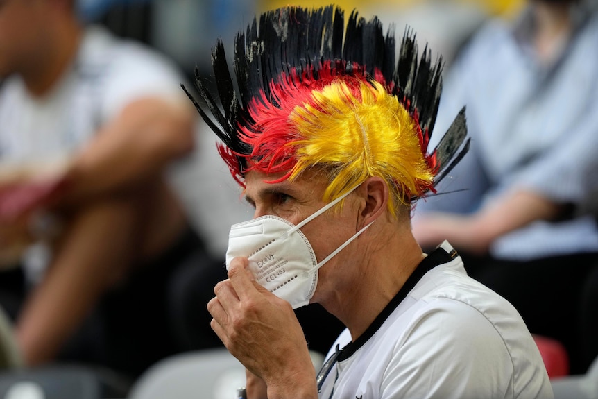 A man with a black, red and yellow mohawk, holds a mask in front of his face
