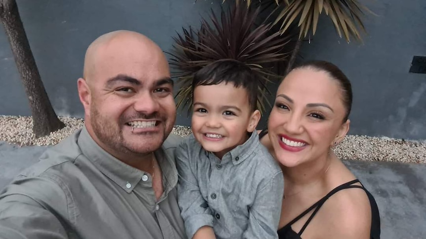 A selfie photo of a family of three: two smiling middle-aged parents and a 3 year old boy in the middle.