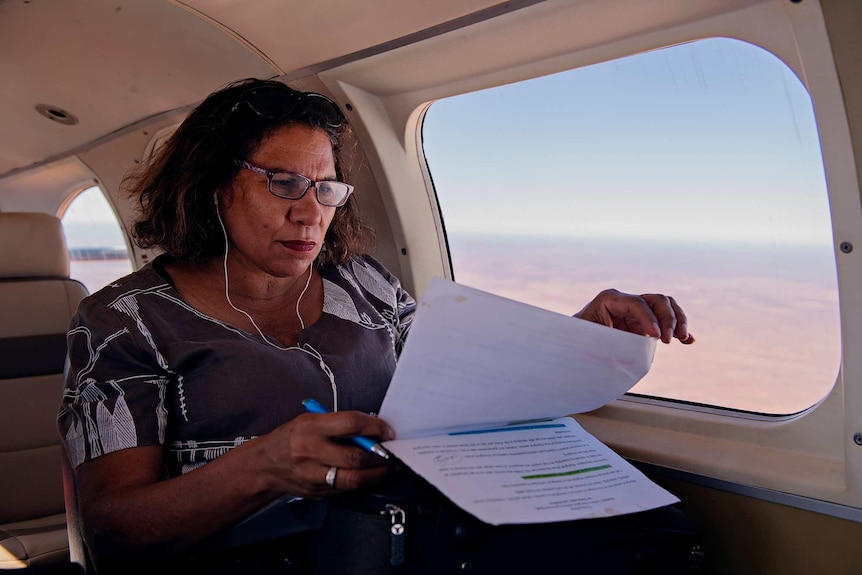 A middle-aged woman wearing glasses looks through documents on plane holding a pen in her hand.