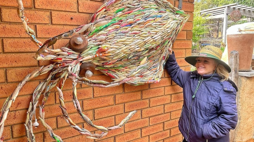 woman with blonde hair in wide-brimmed straw hat and blue waterproof jacket holding squid artwork against brick wall