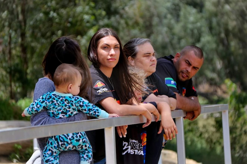 Apryl staring toward the camera, surrounded by family members, with hands over the edge of a bridge barrier, trees behind