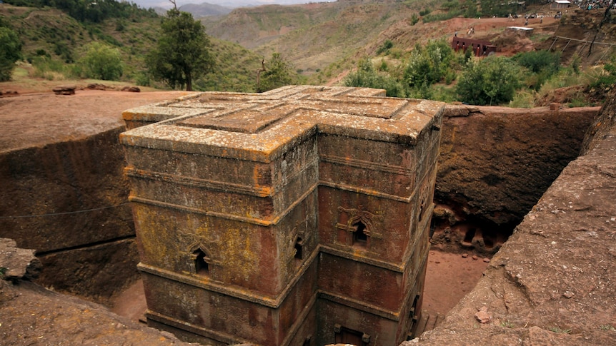 A brown ancient crypt sits in a hole in the ground with rugged canyon behind on a sunny day.