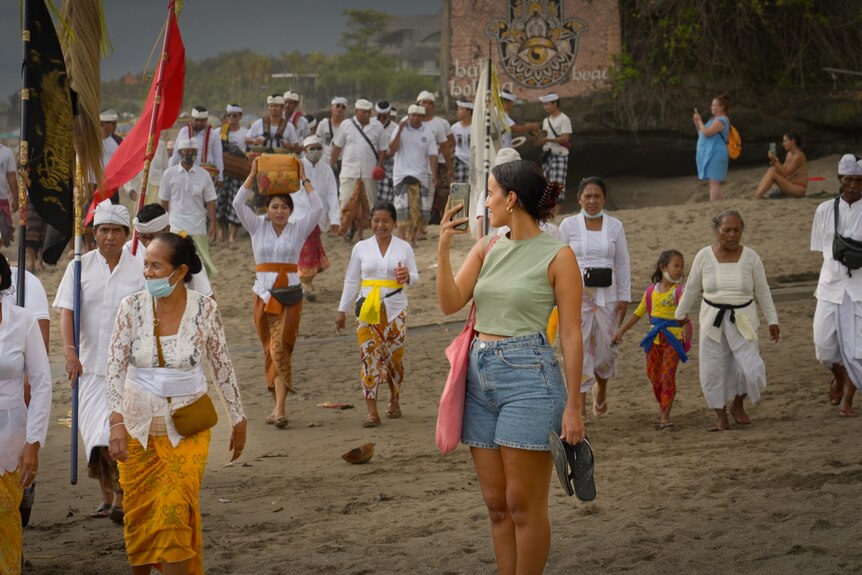 A woman in shorts watches a traditional Balinese parade unfold on a beach 